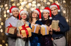 celebration, christmas and holiday concept - happy friends in santa hats with gifts at party over foil fringe and garland lights on background